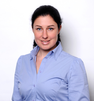Eine Frau mit heller Haut, zurückgebundenem dunklem Haar und blauen Augen lächelt leicht. Sie trägt ein hellblaues Hemd mit offenem Kragen. Der Hintergrund ist schlicht weiß, und sie blickt in professioneller Pose in die Kamera.
