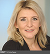 Eine Frau mit schulterlangem blondem Haar und heller Haut lächelt sanft. Sie trägt einen dunklen Blazer über einem hellen Oberteil. Der Hintergrund ist in blassen Blau- und Beigetönen sanft verschwommen. Die gleichmäßige Beleuchtung betont ihre Gesichtszüge.