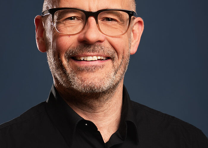 Ein lächelnder Mann mittleren Alters mit Glatze, kurzem Bart und Brille, der ein schwarzes Hemd mit Kragen trägt, posiert vor einem schlichten dunkelblauen Hintergrund.